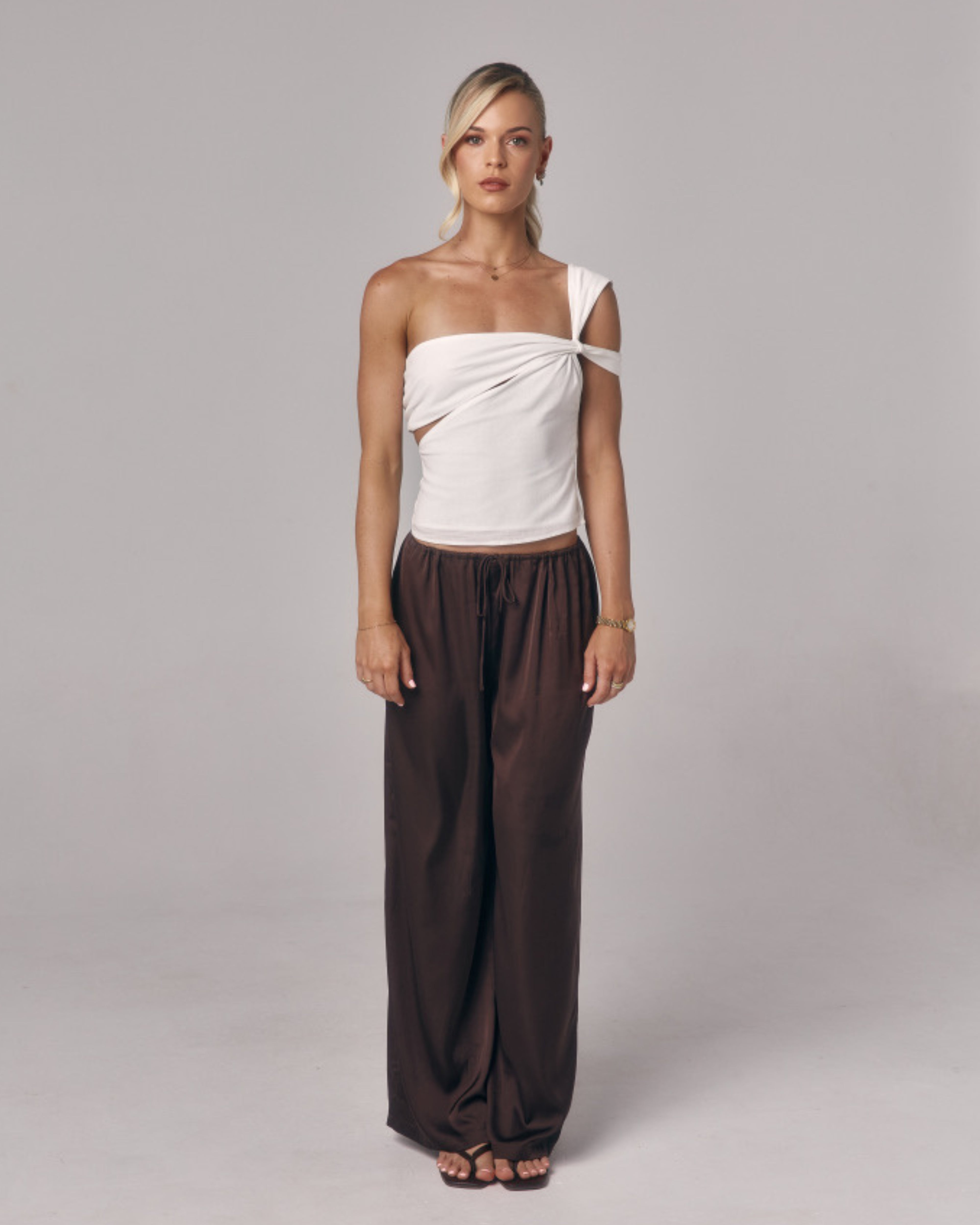 Model wearing Paloma chocolate brown satin lounge pants with drawstring waist styled with a white one shoulder top in studio.
