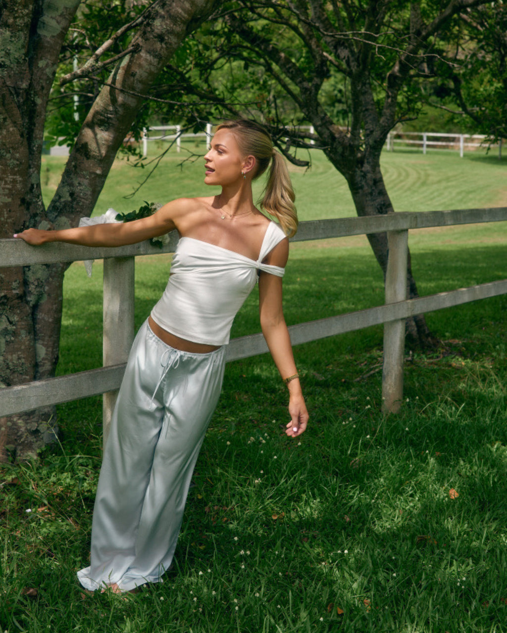 Model wearing Paloma light blue satin pants with matching top outdoors beside a white fence and trees.