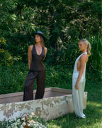 Models wearing Paloma satin lounge pants and matching tops in chocolate brown and light blue in a lush outdoor setting.