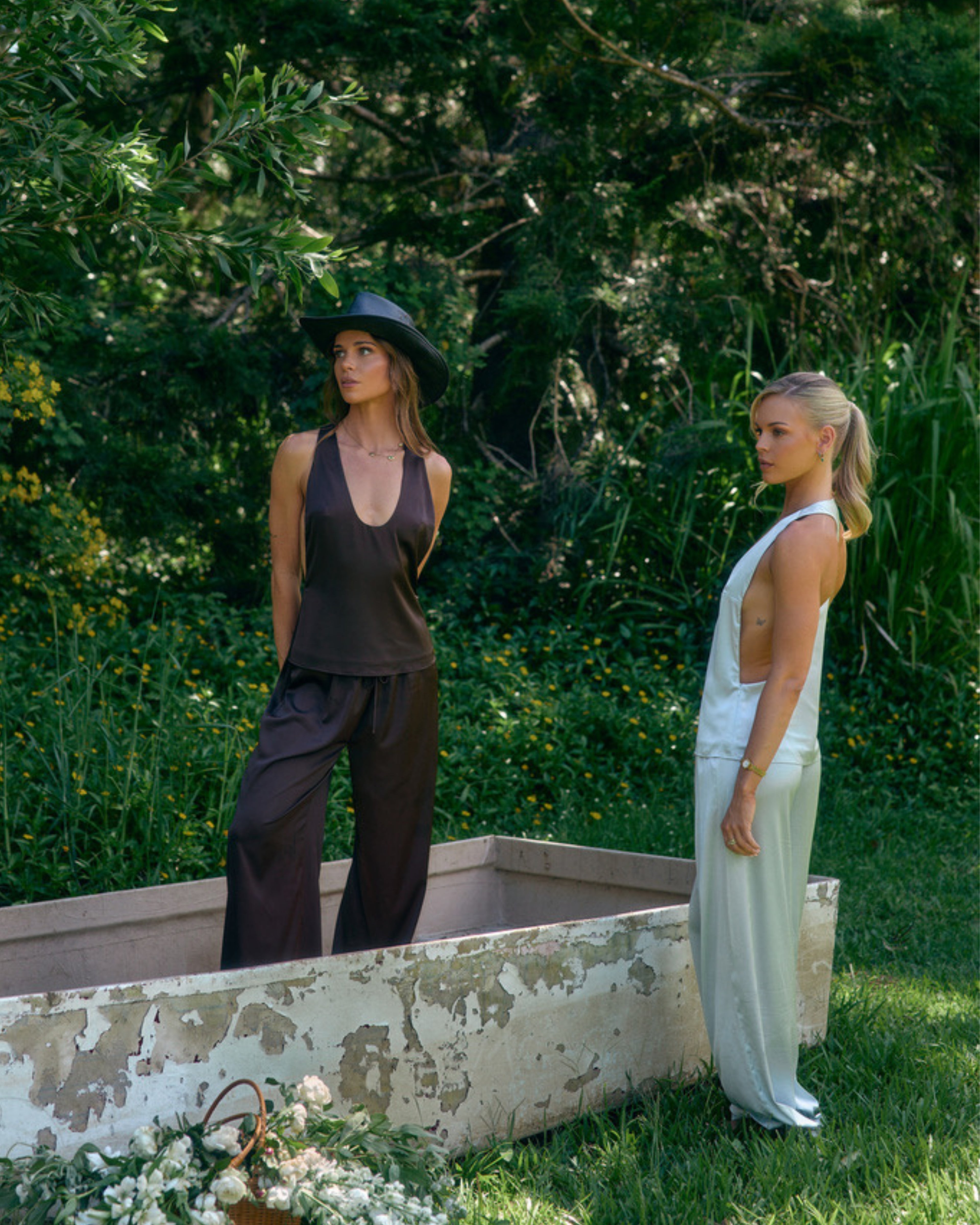 Models wearing Paloma satin lounge pants and matching tops in chocolate brown and light blue in a lush outdoor setting.
