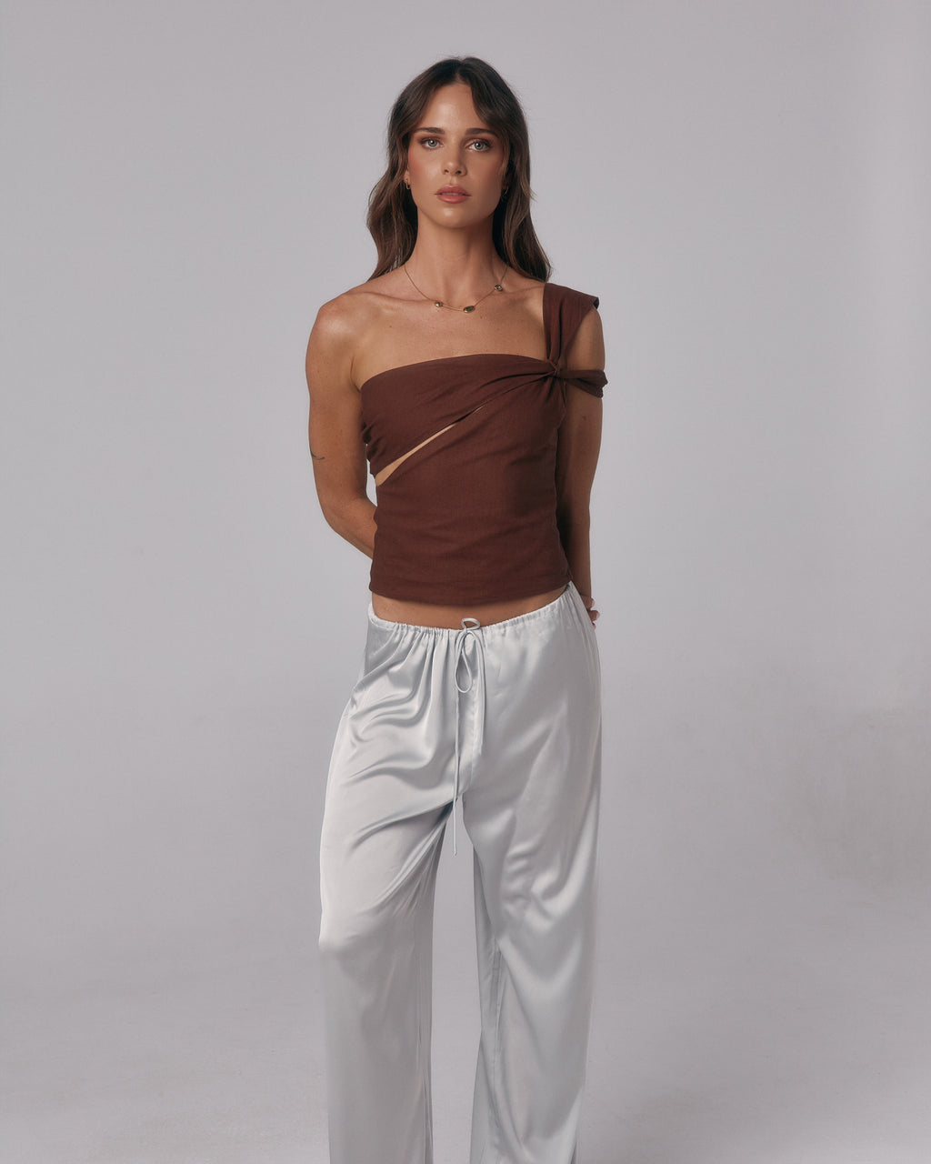 Front view of model wearing the Sienna Top in espresso, an asymmetric one-shoulder fitted top styled with satin pants in studio.