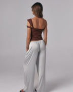 Back view of model wearing the Sienna Top in espresso, an asymmetric off-shoulder fitted top styled with satin pants in studio.