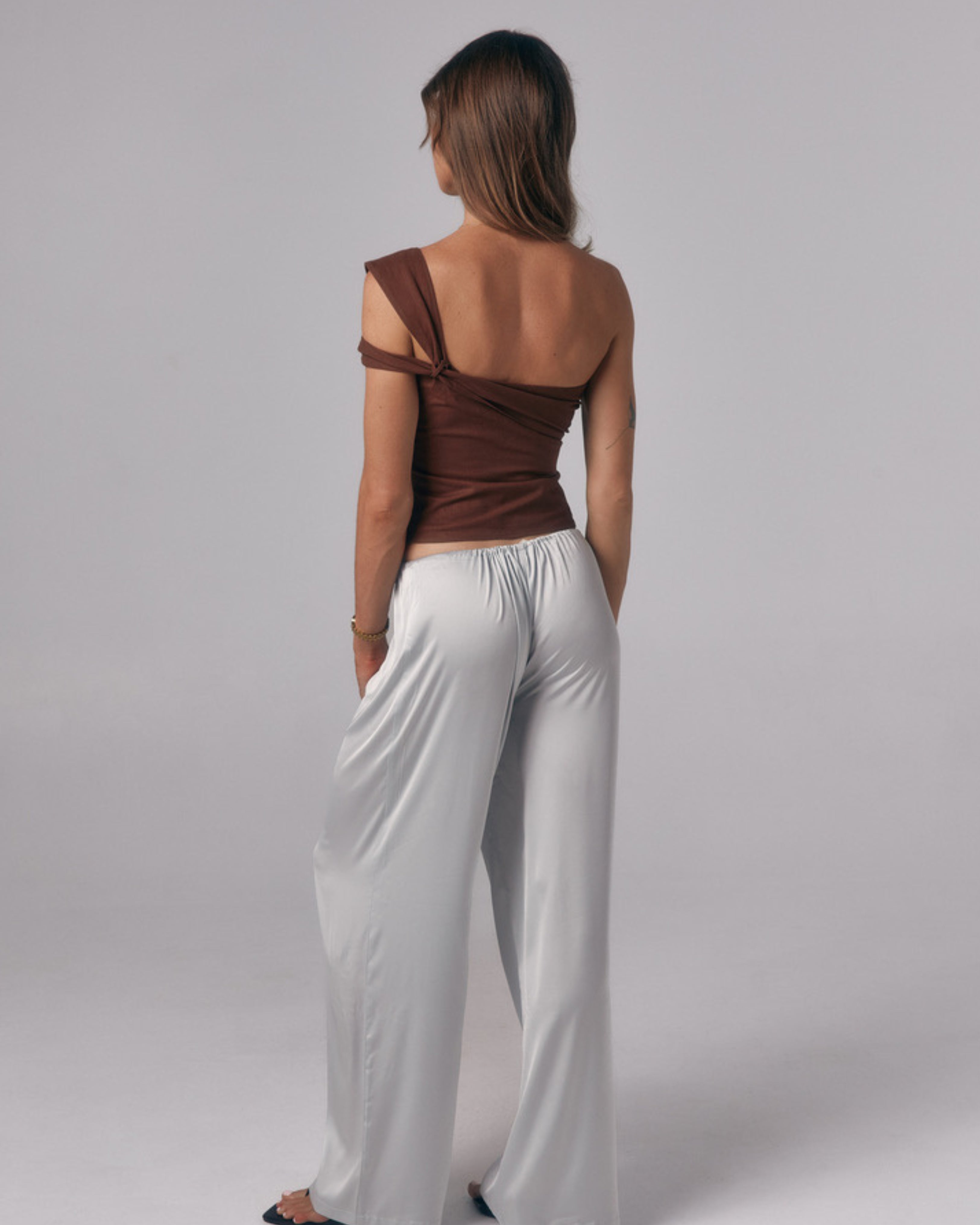 Back view of model wearing the Sienna Top in espresso, an asymmetric off-shoulder fitted top styled with satin pants in studio.