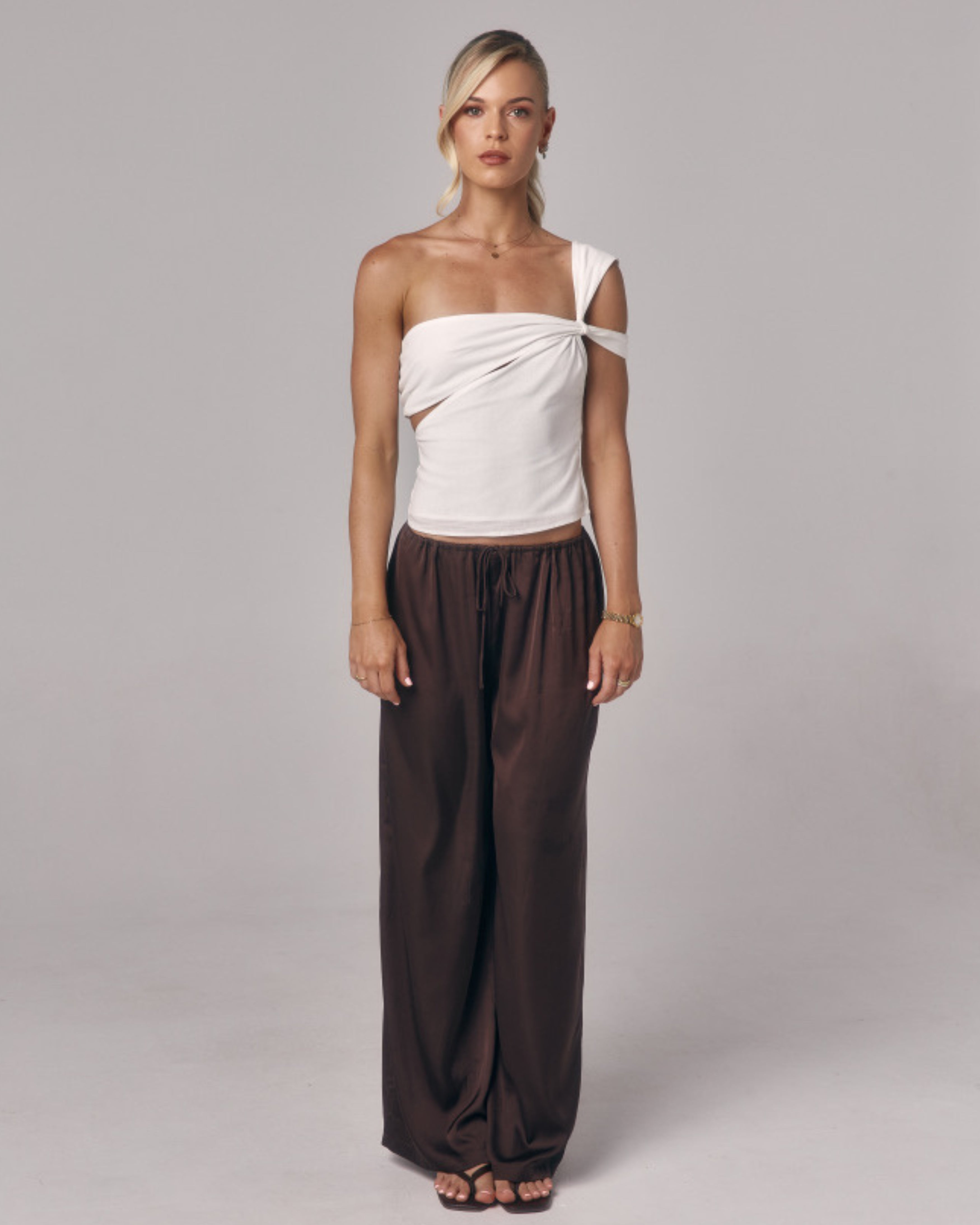 Front view of model wearing the Sienna Top in ivory, an asymmetric one-shoulder fitted top styled with relaxed brown pants in studio.
