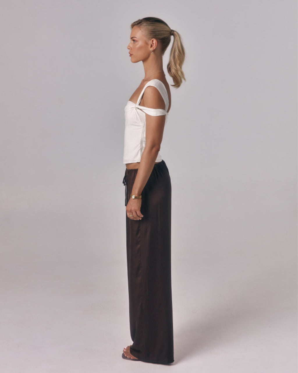 Side view of model wearing the Sienna Top in ivory, an asymmetric off-shoulder fitted top styled with relaxed brown pants in studio.