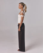Side view of model wearing the Sienna Top in ivory, an asymmetric off-shoulder fitted top styled with relaxed brown pants in studio.