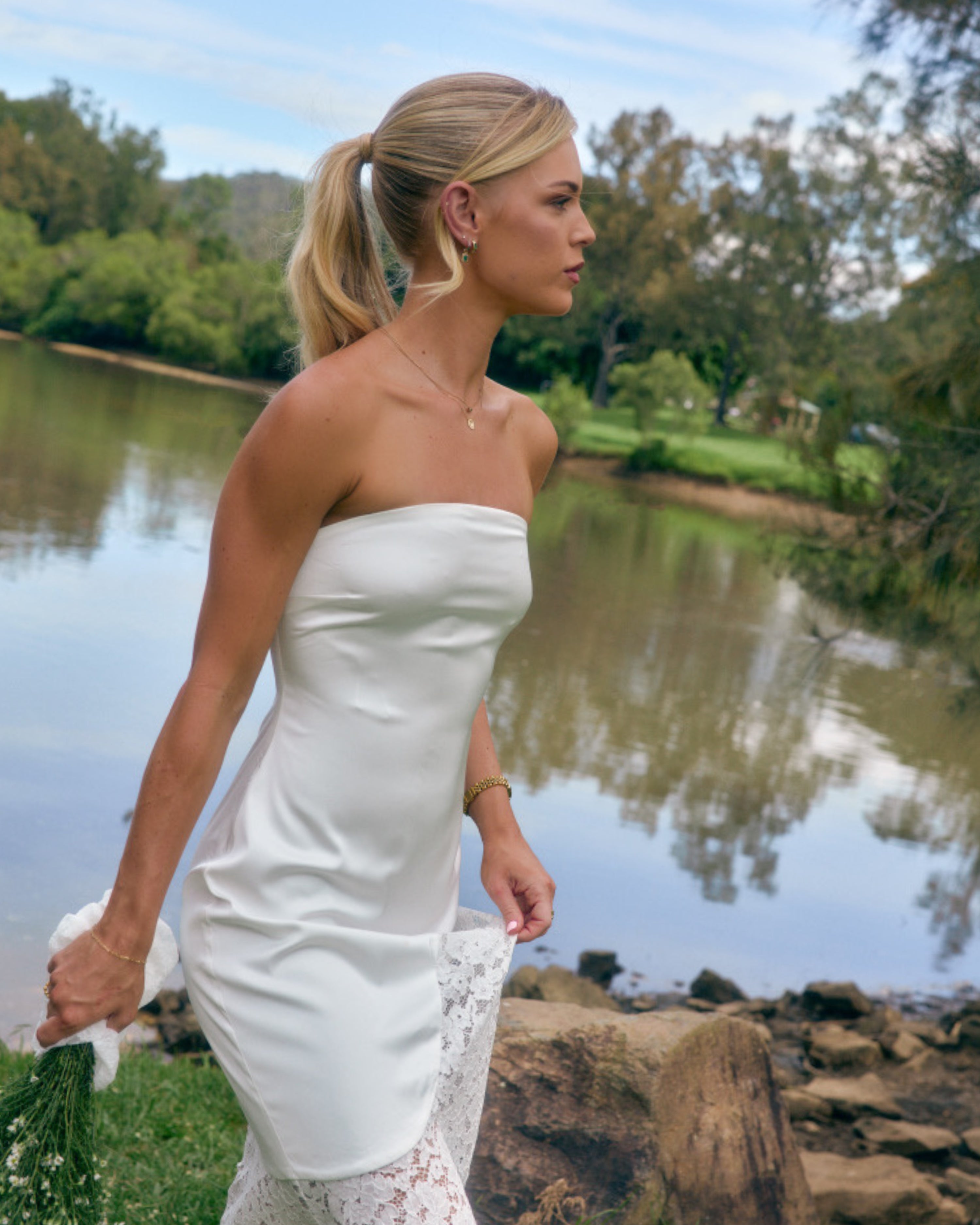 Model wearing Soleil ivory strapless maxi dress with lace skirt panel outdoors beside a lake.